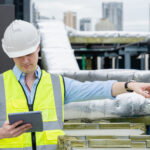 BIM manager in a white hard hat and safety vest checking a tablet while pointing to insulated ductwork and piping