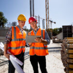 Two construction leaders in hard hats reviewing plans on a tablet at a large data center campus build site with tower cranes
