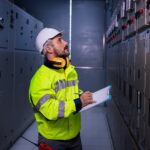 Electrician in a hard hat and hi-vis jacket reviewing a clipboard beside a row of electrical switchgear