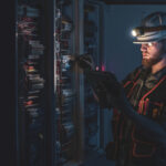 Electrician in a hard hat and headlamp checking an electrical control panel with a tablet