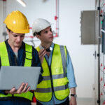 Two data center construction workers in hard hats reviewing project data on a laptop beside electrical switchgear