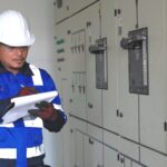 Safety manager in a hard hat and safety vest reviewing a clipboard beside electrical switchgear at a data center construction site