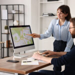 Fiber business development manager pointing to a route map on a computer screen while a colleague reviews it at his desk