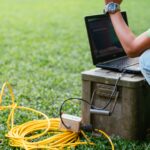 Field technician testing a yellow cable connection with a laptop on a residential lawn during a fiber drop installation