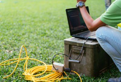 Field technician testing a yellow cable connection with a laptop on a residential lawn during a fiber drop installation