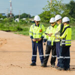 Field crew in hard hats and high-visibility gear reviewing route plans on tablets along a rural construction route
