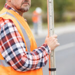 Field worker in an orange safety vest holding a measuring rod along a road while a crew member works in the background