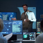Network operations leader briefing his team beside a network monitoring dashboard in an operations center